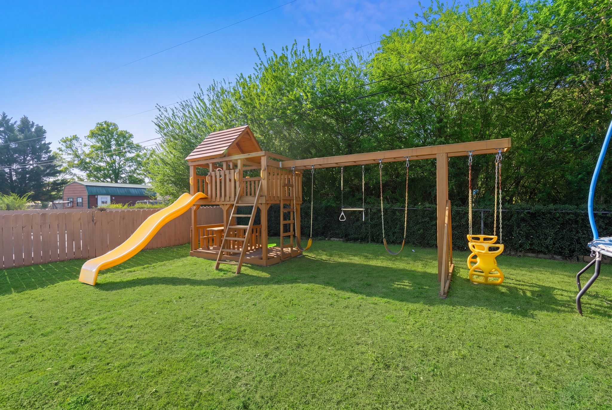 Kids playground area with orange slide, wooden tower, swings, and trampoline on lush green lawn surrounded by mature trees