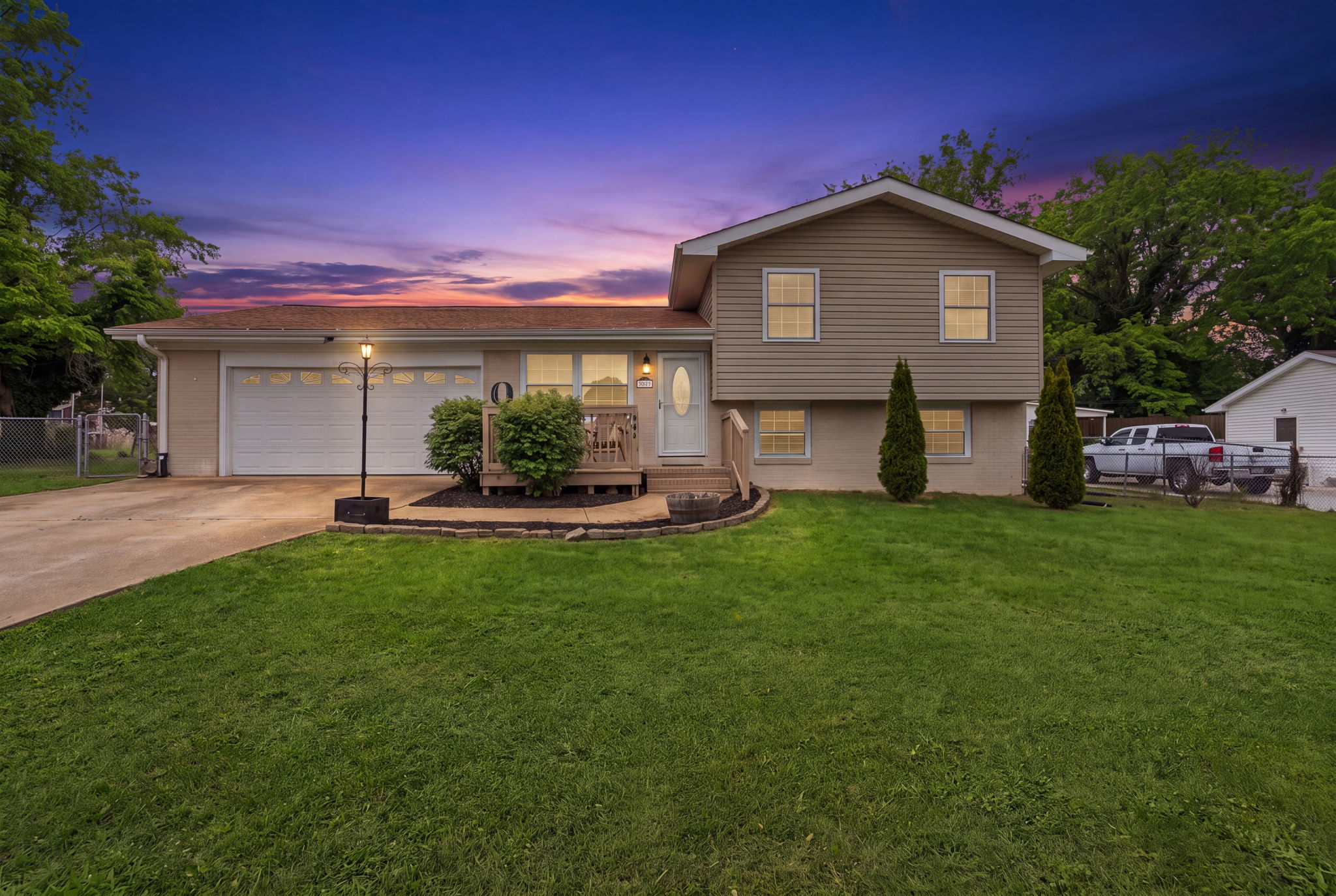 Stunning sunset view of two-car garage home with front porch, manicured lawn, and landscape lighting at dusk